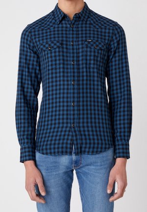 Homme portant une chemise à carreaux bleue et noire à manches longues avec deux poches poitrine et un jean bleu clair.