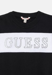 Svart stickad tröja med ett vitt horisontellt rand som har ordet "GUESS" i silverfärgade rhinestones. Ribbad krage och muddar.