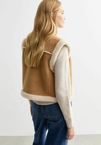 Brun konstläder väst med krämfärgad lammskinnsdetalj, figursydd design och sidöppningar; kombinerad med ljusblå denimjeans.