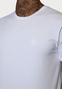 T-shirt en coton blanc avec un col rond, présentant une petite broderie blanche sur la poitrine. Texture lisse, manches courtes.