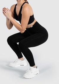 Kobieta w czarnym stroju sportowym i białych sneakersach wykonująca ćwiczenie przysiadu na jednolitym jasnym tle.