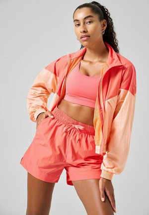 Chaqueta cortavientos de color coral con puños elásticos, combinada con un sujetador deportivo y pantalones cortos a juego en color coral. Tejido ligero y texturizado, ajuste casual.