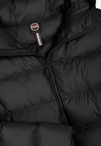 Melnā pūkainā jaka ar augstu apkakli, kvadrātveida dizainu un pilnu rāvējslēdzēju. Iekļauta markas birka ar sarkaniem, baltiem un pelēkiem akcentiem.