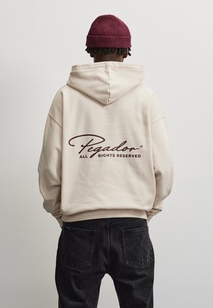 Personne portant un sweat à capuche beige avec le texte "Pegador All Rights Reserved" dans le dos et un bonnet tricoté bordeaux, debout sur un fond uni.