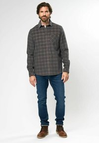 Camisa de cuadros gris oscuro con botones, que presenta un bolsillo en el pecho y mangas largas, combinada con jeans azules y botas de cordones marrones.