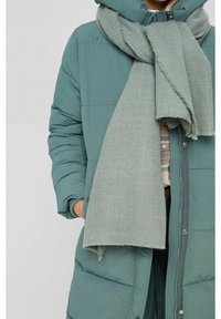 Personne portant un manteau matelassé bleu sarcelle avec les mains dans les poches et une grosse écharpe gris clair enroulée autour du cou.