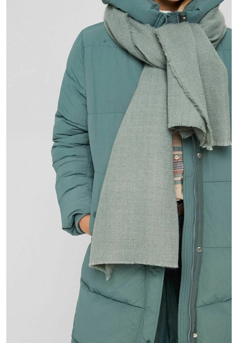 Personne portant un manteau matelassé bleu sarcelle avec les mains dans les poches et une grosse écharpe gris clair enroulée autour du cou.