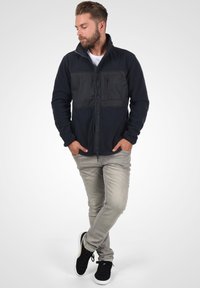 Chaqueta de forro polar en azul marino con paneles negros, con cremallera completa y bolsillo en el pecho. Combinada con jeans gris claro y zapatillas negras.