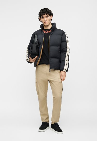 Ung mand iført sort dunjakke med hvide striber, sort skjorte, beige cargobukser og sorte sneakers, der holder en sort håndtaske.