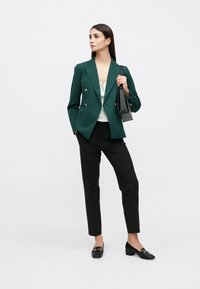 Blazer vert croisé, haut blanc, pantalon noir, et mocassins noirs avec accents dorés. Tissu lisse, coupe ajustée, design minimaliste.