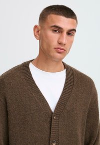 Brown knitted cardigan with a V-neck, ribbed edges, and wooden buttons. Worn over a white t-shirt. Soft texture, relaxed fit.