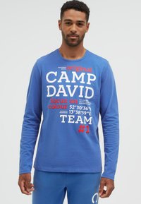 Mann trägt ein blaues Langarmshirt mit weißem und rotem "CAMP DAVID"-Schriftzug und Koordinaten, kombiniert mit passenden blauen Hosen.