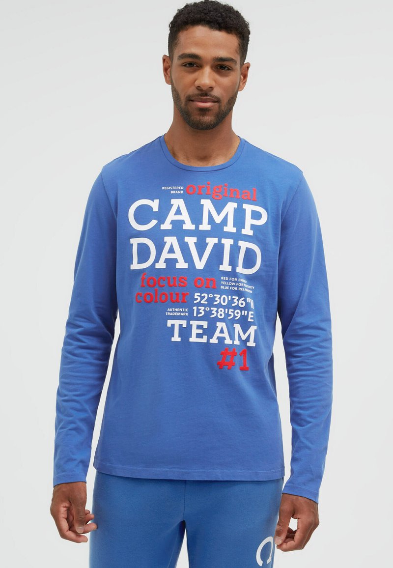 Mann trägt ein blaues Langarmshirt mit weißem und rotem "CAMP DAVID"-Schriftzug und Koordinaten, kombiniert mit passenden blauen Hosen.