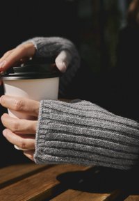 Grijze geribde, gebreide vingerloze handschoenen houden een witte beker met een zwart deksel vast, met een zachte textuur en subtiele glans tegen een houten oppervlak.