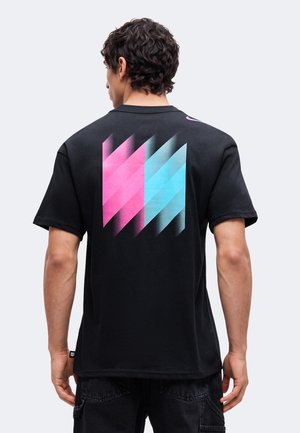 Homme aux cheveux bouclés et foncés, portant un T-shirt noir avec des formes géométriques roses et bleues dans le dos, debout devant un fond uni.