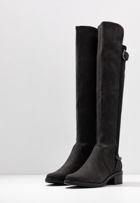 Botas negras hasta la rodilla, hechas de material similar al ante, con un acabado suave, punta redondeada y detalles con hebillas en los laterales. Adecuadas para el uso invernal.