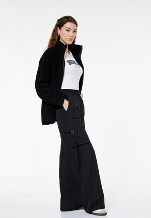 Kobieta ubrana w czarną polarową kurtkę, białą koszulkę z nadrukiem i czarne szerokie spodnie cargo, stoi z rękami w kieszeniach.