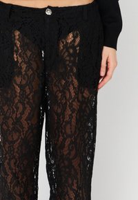 Pantaloni neri in pizzo caratterizzati da un design trasparente con motivi floreali. Chiusura in vita con un bottone argento. Testurizzati con dettagli intricati.