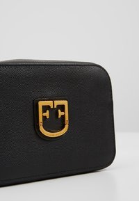 Sort tekstureret læderhåndtaske med en guldfarvet prægning af logoet på låsen foran, placeret mod en ensfarvet grå baggrund.