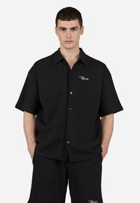Black short-sleeve shirt featuring a button-up front, pointed collar, and textured fabric. White logo accent on the left chest area.