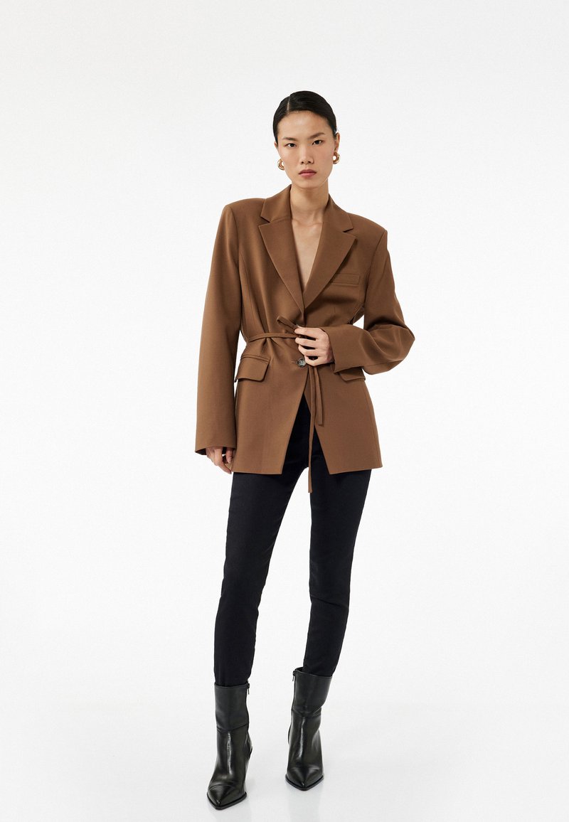 Blazer marrón a medida con cintura ceñida, solapas de pico y bolsillos de solapa, combinado con vaqueros skinny negros y botas de tacón negras.