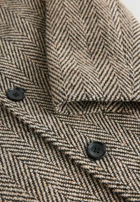 Herringbone-mønsteret stof i beige og sort, med en blød uldtekstur og en sort knapdetalje på venstre side.