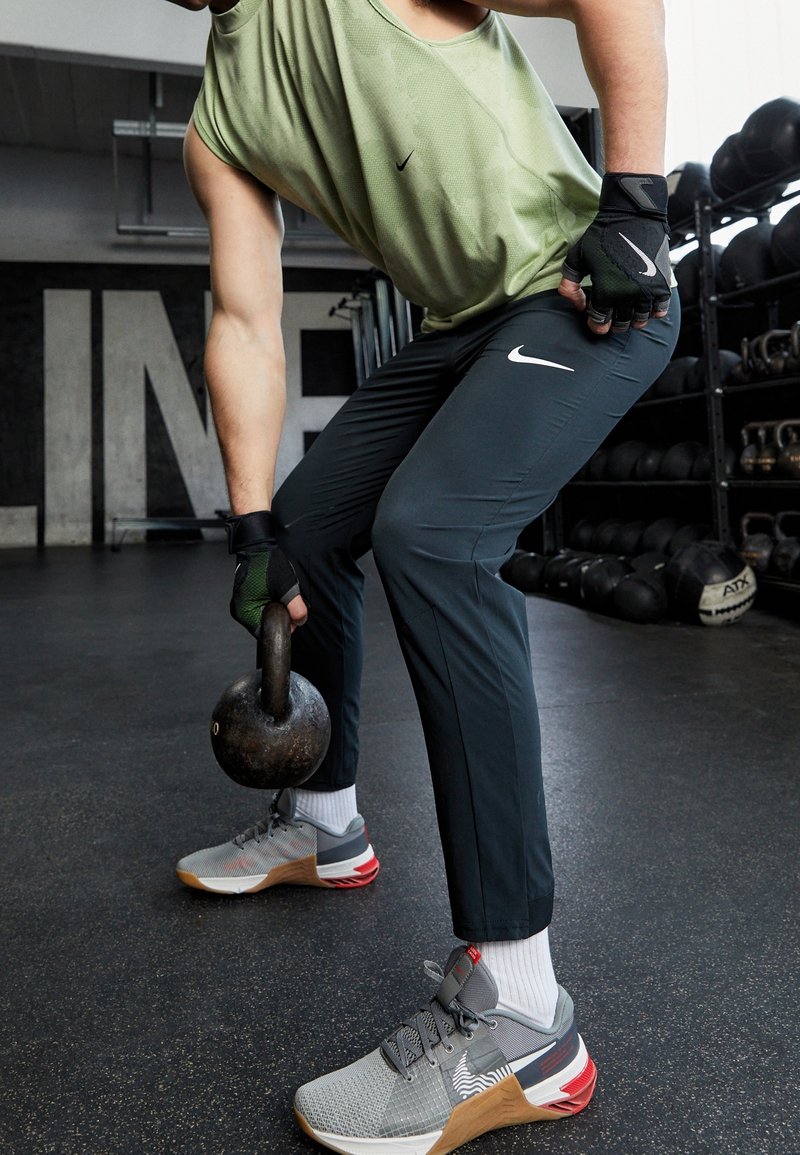 Šedé sportovní boty s červenými akcenty, tmavé flexibilní kalhoty a světle zelený top bez rukávů. Model zvedá kettlebell.