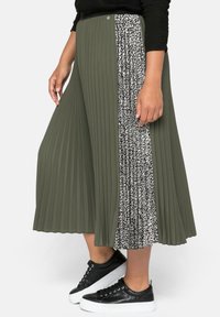 Jupe plissée vert olive avec un panneau latéral au motif léopard noir et blanc, assortie à des baskets noires.
