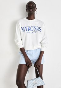 Hvit hettegenser med blå tekst "MYKONOS BLUE OCEAN", kombinert med lyseblå shorts, holder en mønstret blå handbag.