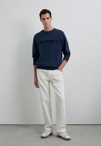 Hombre vestido con un sudadera azul marino con el texto "scalpers", pantalones rectos blancos y zapatillas blancas, de pie contra una pared lisa.