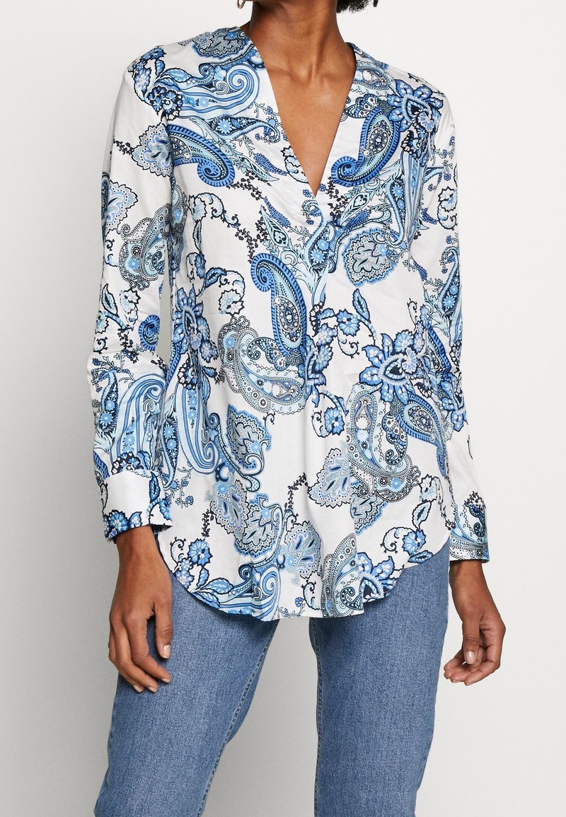 Blouse blanche à manches longues avec motif floral paisley bleu, portée avec un jean bleu clair, avec un décolleté en V profond et des poignets boutonnés.