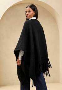 Poncho nero con texture, con bordi frangiati, indossato sopra una maglietta bianca. Vestibilità ampia, realizzato in tessuto morbido, con una silhouette drappeggiata.