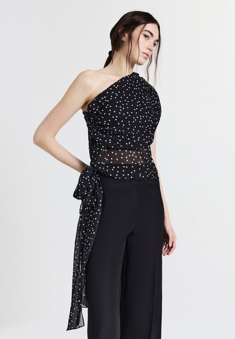 Femme portant un haut à pois noir à une épaule avec une section mi-transparent et un pantalon large noir devant un fond uni.