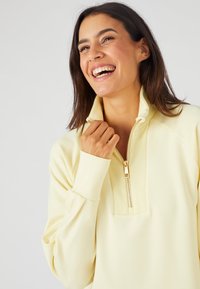 Un suéter amarillo claro con cuello con cremallera, detalles acanalados en el escote y tejido suave. Presenta mangas largas y un corte relajado.