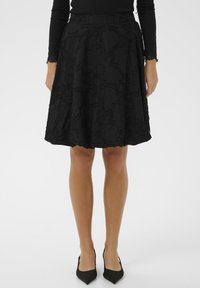 Ženska v črni strukturirani A-linijski krili, oblečena v prilagojen črn dolg rokav in črne čevlje s koničastim nosom, stoji ob enobarvni ozadju.