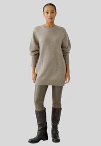Beiger oversized Pullover mit gerippten Bündchen und Saum, kombiniert mit olivgrünen Lederleggings und schwarzen kniehohen Stiefeln mit Schnallen-Detail.