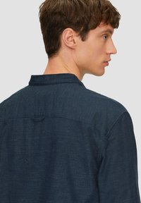 Chemise bleu marine en tissu texturé, avec un col court et un détail de rainure au dos. Coupe lisse et ajustée avec des accents de couture subtils.