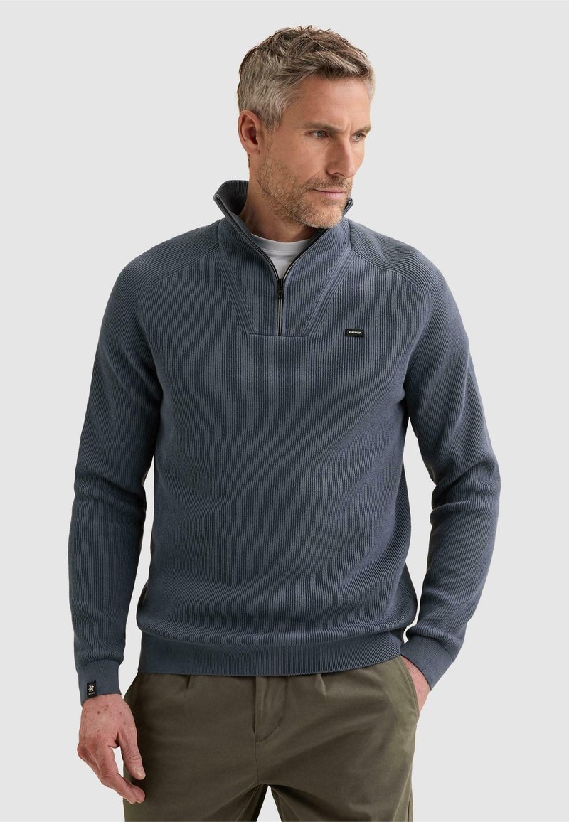 Homme d'âge moyen aux cheveux gris, portant un pull bleu à demi-fermeture éclair et un pantalon vert olive, debout avec une main dans la poche devant un fond uni.