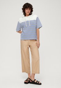 Gestreifter blauer und weißer Kapuzenpullover kombiniert mit beigen weit geschnittenen Hosen und schwarzen Sandalen. Der Pullover hat eine kontrastierende Passe und eine Kordelzug.