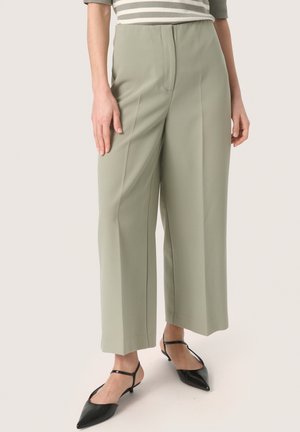 Helle grüne weite Hose mit hoher Taille, die eine glatte Textur und subtile Seitennahtdetails aufweist, kombiniert mit schwarzen spitzen Schuhen.