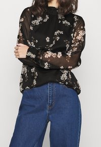 Blouse noire à fleurs en tissu transparent avec manches longues bouffantes, poignets élastiques et encolure froncée, associée à un jean en denim bleu.