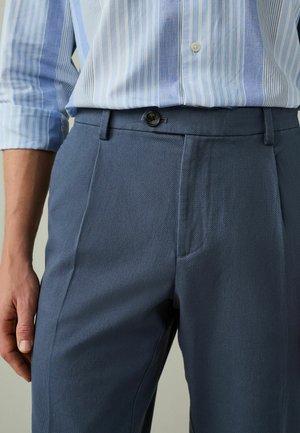 Man wearing blue textured dress pants and a light blue striped button-up shirt with sleeves rolled up.