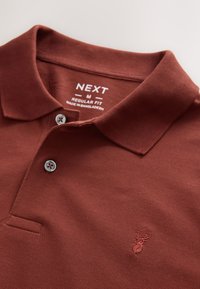 Polo marrón con dos botones, logo de ciervo bordado y etiqueta con la palabra "NEXT", ajuste regular medio, fabricado en Bangladesh.