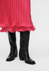 Fekete térdig érő bőrcsizmák blokk sarkakkal, világos rózsaszín pliszírozott midi szoknya alatt, fodros szegéllyel.