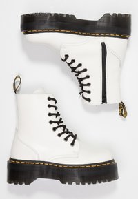 Botas de tobillo de cuero blanco con cordones negros, suela gruesa negra, costuras amarillas, cremallera lateral y tira de tiro en el talón.