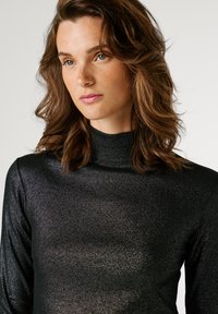 Γυναίκα με κυματιστά καστανά μαλλιά και φακίδες, φορώντας ένα αστραφτερό μαύρο πουλόβερ με ψηλό λαιμό, κοιτάζει ελαφρώς δεξιά σε ένα απλό ανοιχτό φόντο.