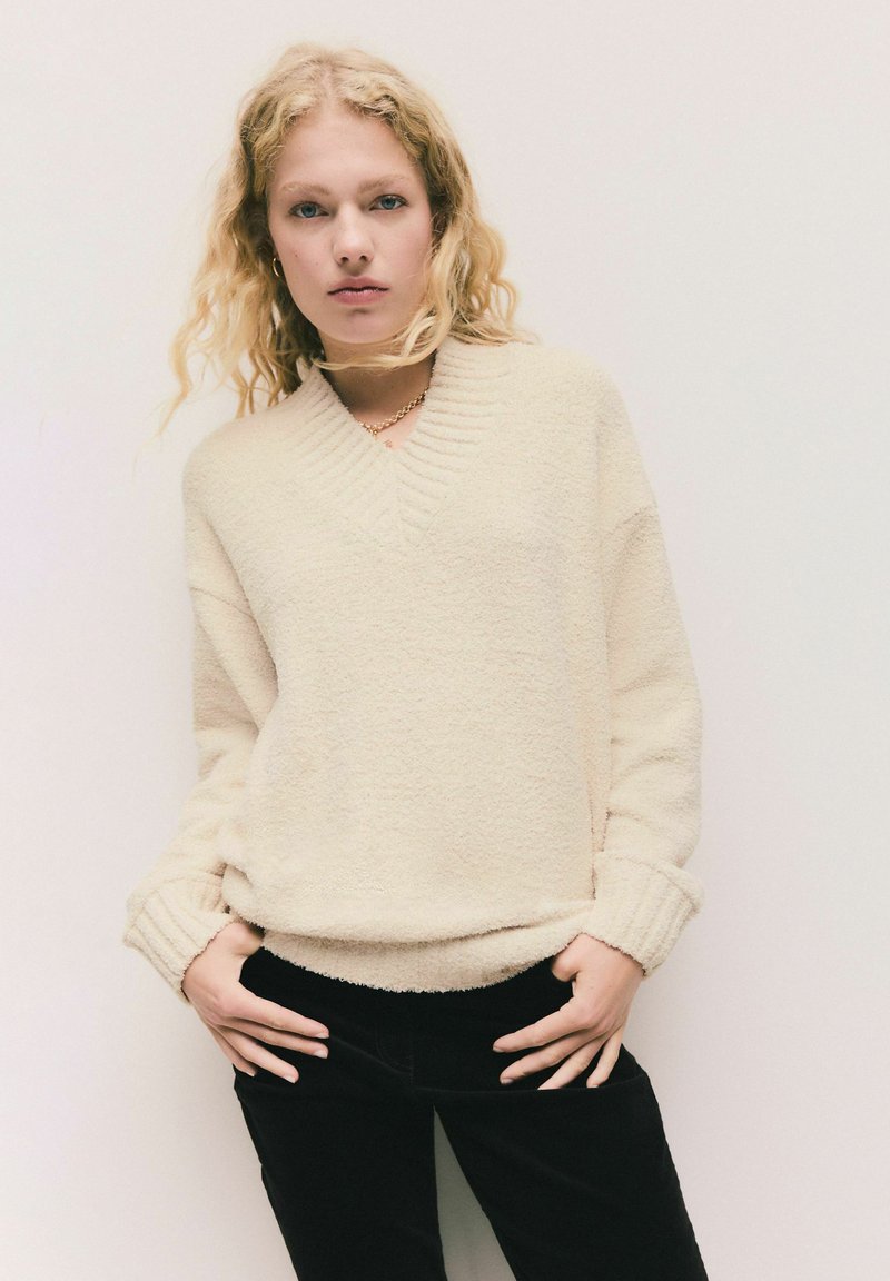 Krémový, texturovaný pletený svetr s výstřihem do V a ribbenými manžetami. Kombinován s černými kalhotami, které mají uvolněný střih a jemný materiál.