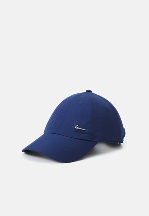 Casquette de baseball bleu marine avec une visière courbée et un petit logo métallique Nike swoosh à l'avant, posée sur une surface blanche.