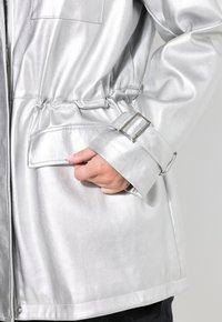 Hopeat metalliväri takki, jossa on kiristettävä vyötärö, solkidetalji hihassa ja etutasku. Väri sileä pinta ja tyylikäs muotoilu.