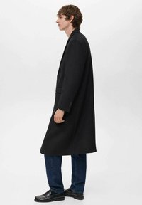 Joven de perfil vestido con un abrigo largo negro, jeans azules y mocasines de cuero negro sobre un fondo blanco liso.